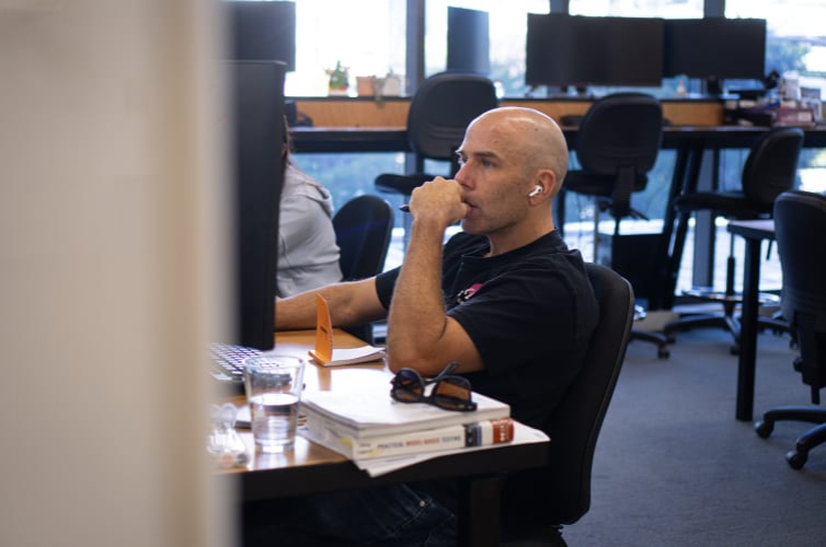 A person working in a modern office, focused on a computer screen with books and a glass of water nearby. The individual is seated in a collaborative workspace, wearing AirPods and deep in thought, reflecting a tech-savvy and productive environment.