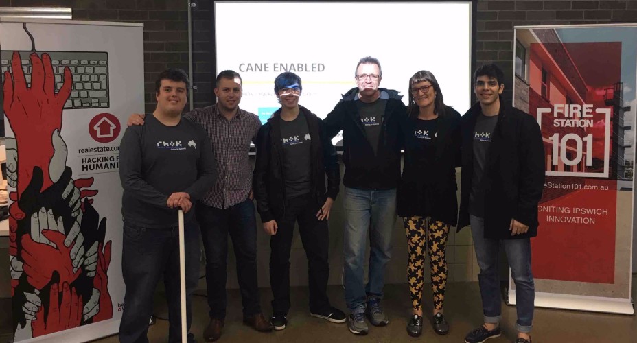 Group photo of the team behind the ‘Cane Enabled’ project, standing in front of a presentation screen. The team members are smiling and wearing RHoK t-shirts, showcasing their success in a hackathon event. Two banners are visible, one promoting Fire Station 101, a local innovation hub, and the other representing the hackathon’s theme, ‘Hacking for Humanity